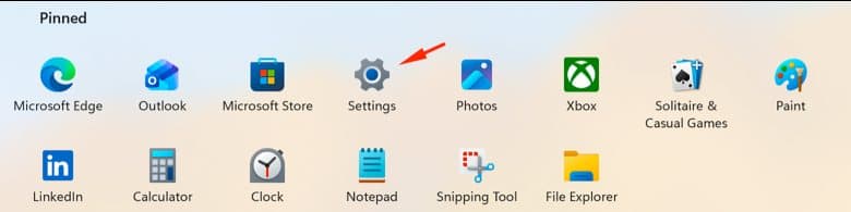 The Windows 11 Start menu pinned apps with an arrow pointing at the Settings gear icon.